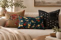 Three decorative throw pillows arranged on a light beige sofa in a styled living room. The center pillow is a dark teal rectangle with a multicolor floral print, flanked by a tan floral pillow and a black patchwork-style pillow with white floral motifs.