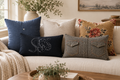 Five decorative throw pillows arranged on a light beige sofa in a styled living room. The pillows include a navy blue pocket-style pillow, a black embroidered pillow, a silver pillow, a western pillow, and a mustard floral pillow.