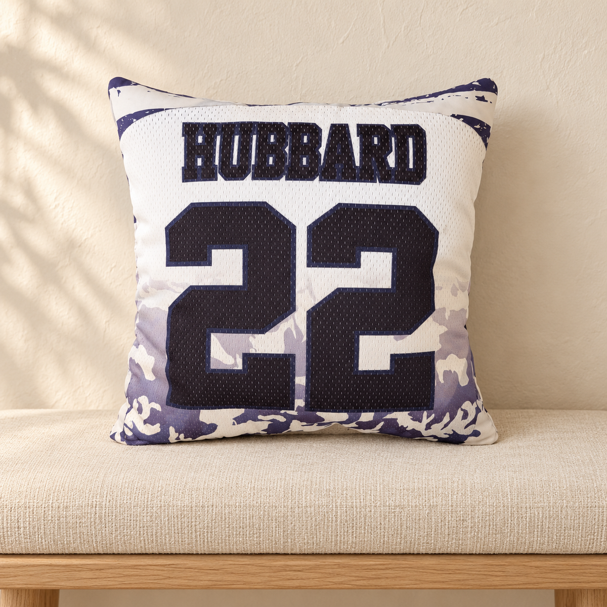 A square throw pillow styled like a sports jersey, featuring the name and number '22' in black on a white mesh background, with a purple and white camouflage pattern along the bottom border, resting on a beige upholstered bench.