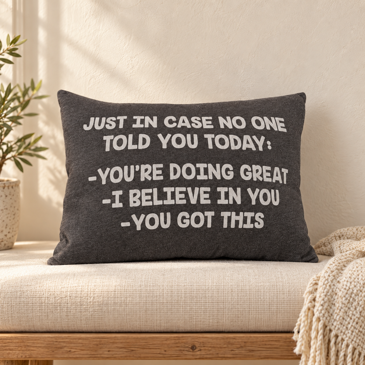 A dark gray rectangular throw pillow with white embroidered text reading 'JUST IN CASE NO ONE TOLD YOU TODAY: -YOU'RE DOING GREAT -I BELIEVE IN YOU -YOU GOT THIS' resting on a beige upholstered bench in a softly lit room with a plant and blanket nearby.