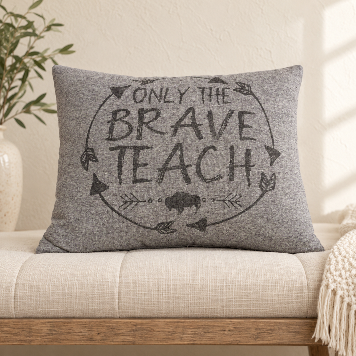 A rectangular gray throw pillow with a dark gray graphic that reads 'ONLY THE BRAVE TEACH' inside a circle with arrows and a buffalo silhouette, resting on a beige upholstered bench in a softly lit room.